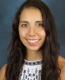 Alyssa Guadagni, with long brown hair and a patterned white top, smiles at the camera against a plain blue background.
