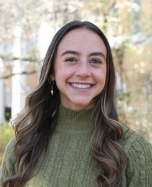 A young woman with long brown hair, wearing a green knit sweater, smiles at the camera outdoors with trees in the background, celebrating her results in the 2024 Graduate Student Research Competition.