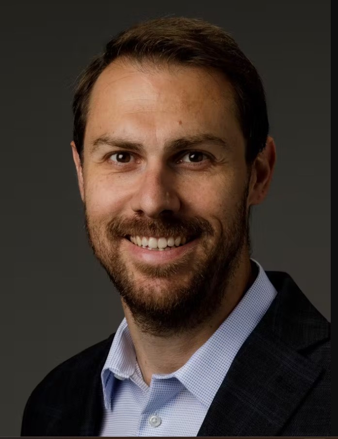 A man with short brown hair and a beard, wearing a dark suit jacket and light blue shirt, smiles at the camera against a dark background—capturing the confident presence of future keynote speakers for the 2026 Symposium.