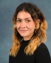 A woman with wavy brown and blonde hair, wearing a black top, is posing in front of a plain blue-gray background, representing a leading research initiative in the study of obesity.