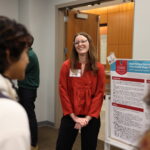 A woman stands beside a research poster, discussing her graduate student research with attendees at an indoor academic event.