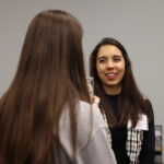 Two women stand indoors having a conversation; one is facing the camera and smiling while discussing graduate student research, as the other has her back to the camera.