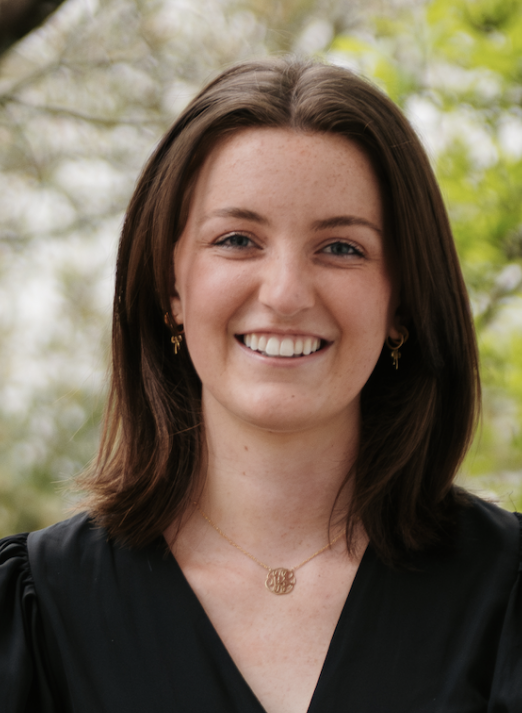A woman with shoulder-length brown hair, wearing a black puff-sleeve dress, stands outdoors in front of green trees, smiling at the camera after the 2024 Research Competition.