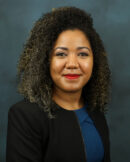 Minaya, Dulce Woman with curly hair, Dulce Minaya, wearing a black blazer and blue top, smiling in front of a plain dark blue background.